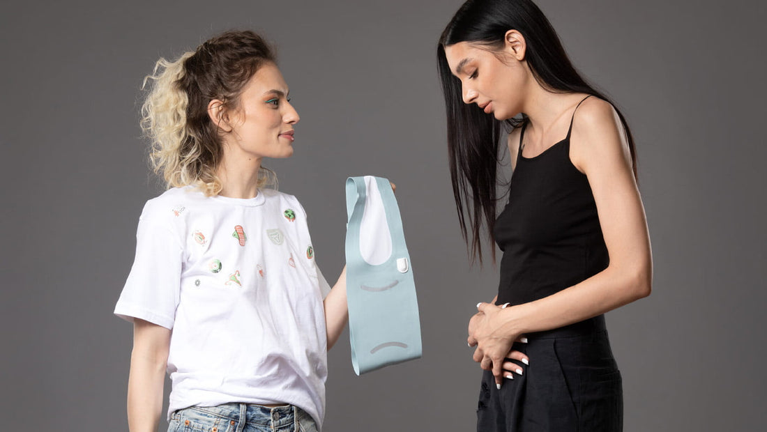 Femeie oferind cu grijă o pungă reutilizabilă unei alte femei care ține mâna pe abdomen, sugerând disconfort menstrual