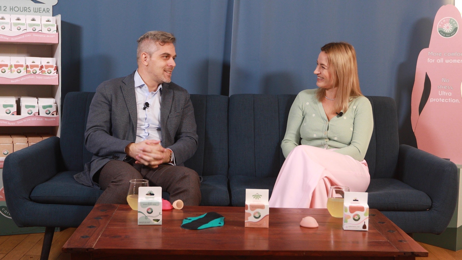 Interviu despre produse menstruale: două persoane discută pe canapea, cu discuri și produse de igienă feminină expuse.