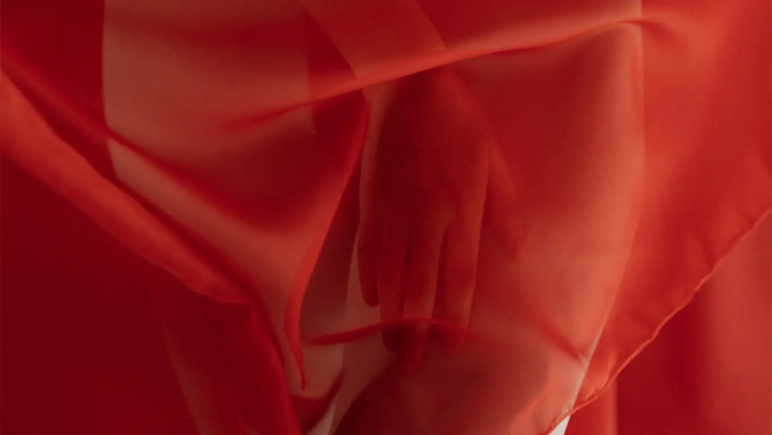 Mână feminină acoperită de un voal roșu translucid – simbol al intimității, feminității și al subiectelor delicate precum menstruația.