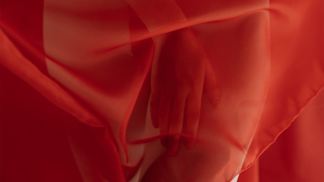 Mână feminină acoperită de un voal roșu translucid – simbol al intimității, feminității și al subiectelor delicate precum menstruația.
